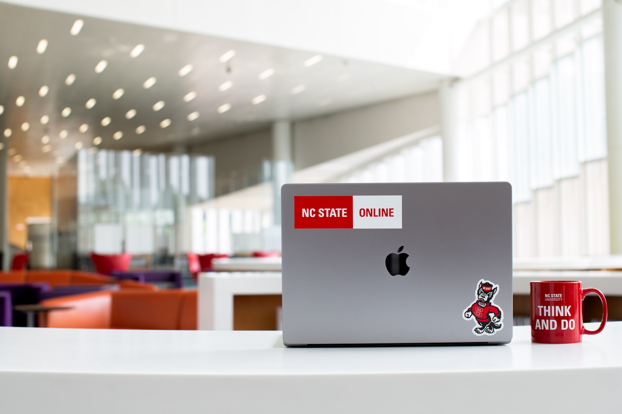 Think and Do coffee mug and a Laptop with NC State Online sticker sit on a table at Hunt Library.