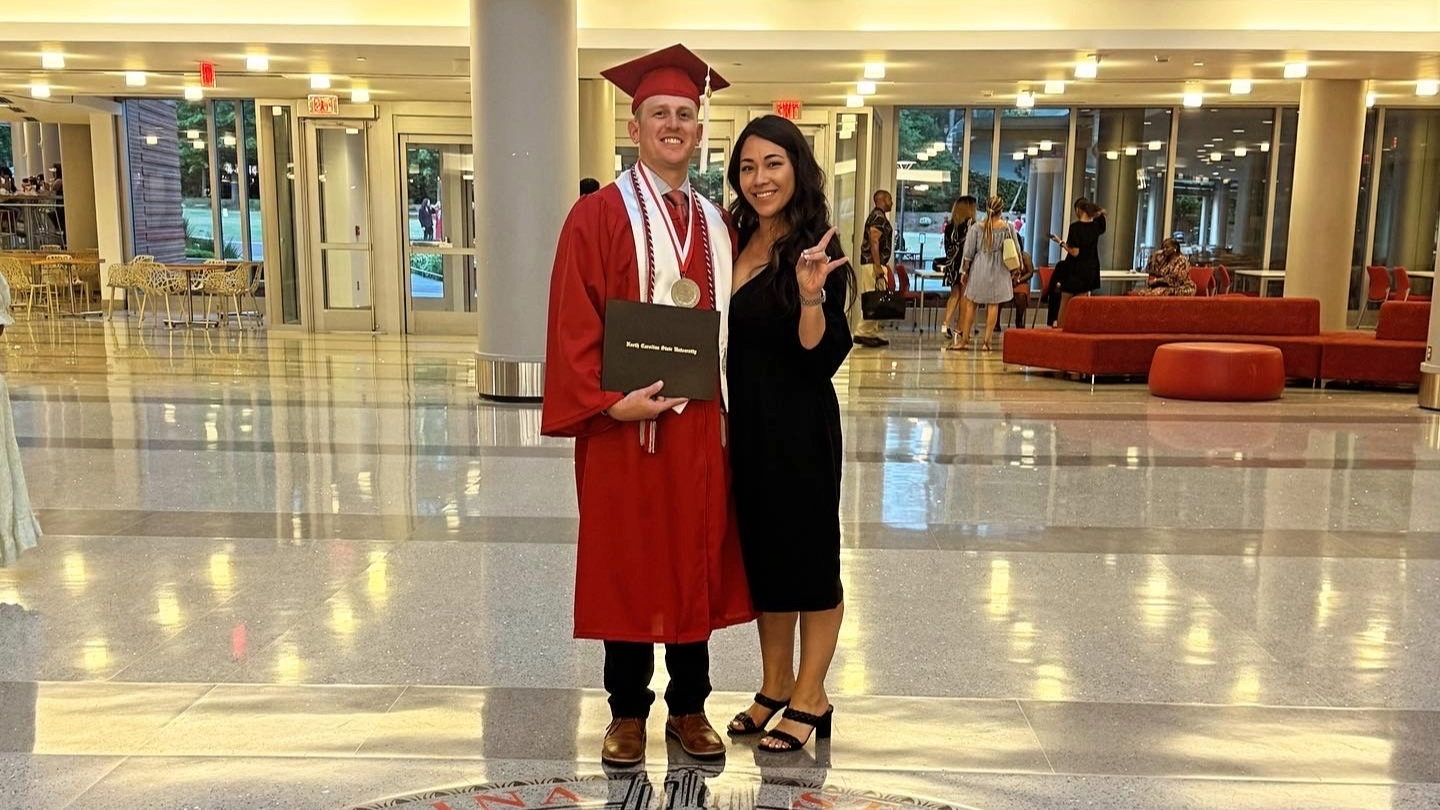 Two people standing in an atrium. The man is in graduation reglaia.