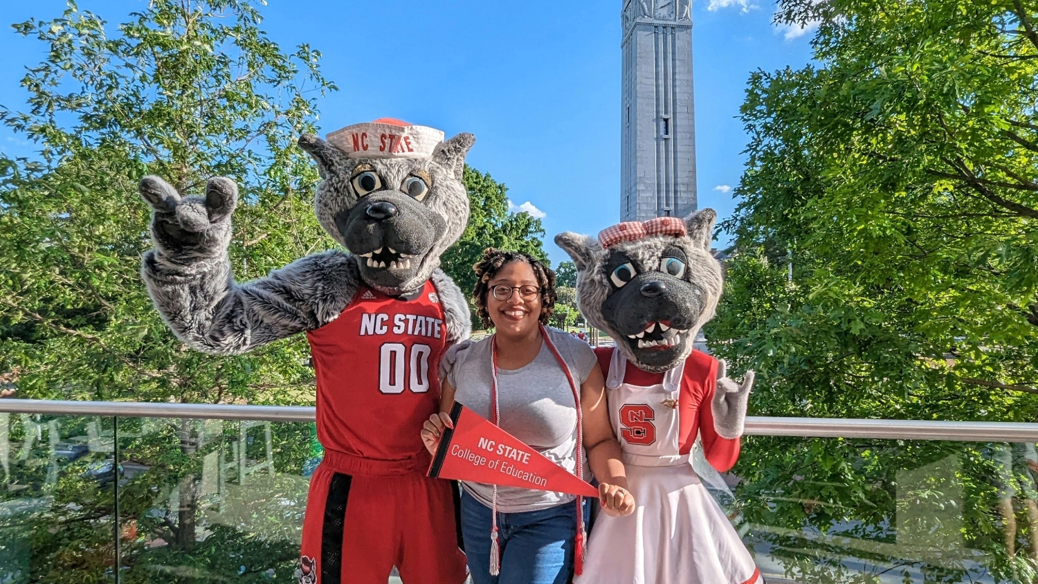 A woman smiling between two NC State mascots outdoors with the NC State belltower in the background.