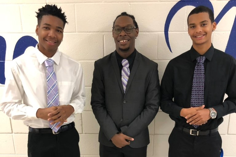 Three men pose in a high school corridor in suits and ties