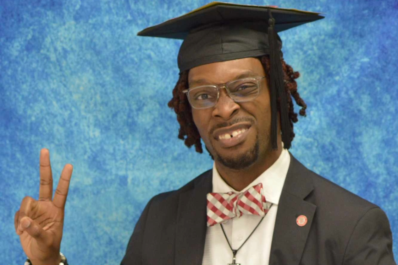 A headshot of a man in graduation regalia holding up a peace sign and making a funny face.